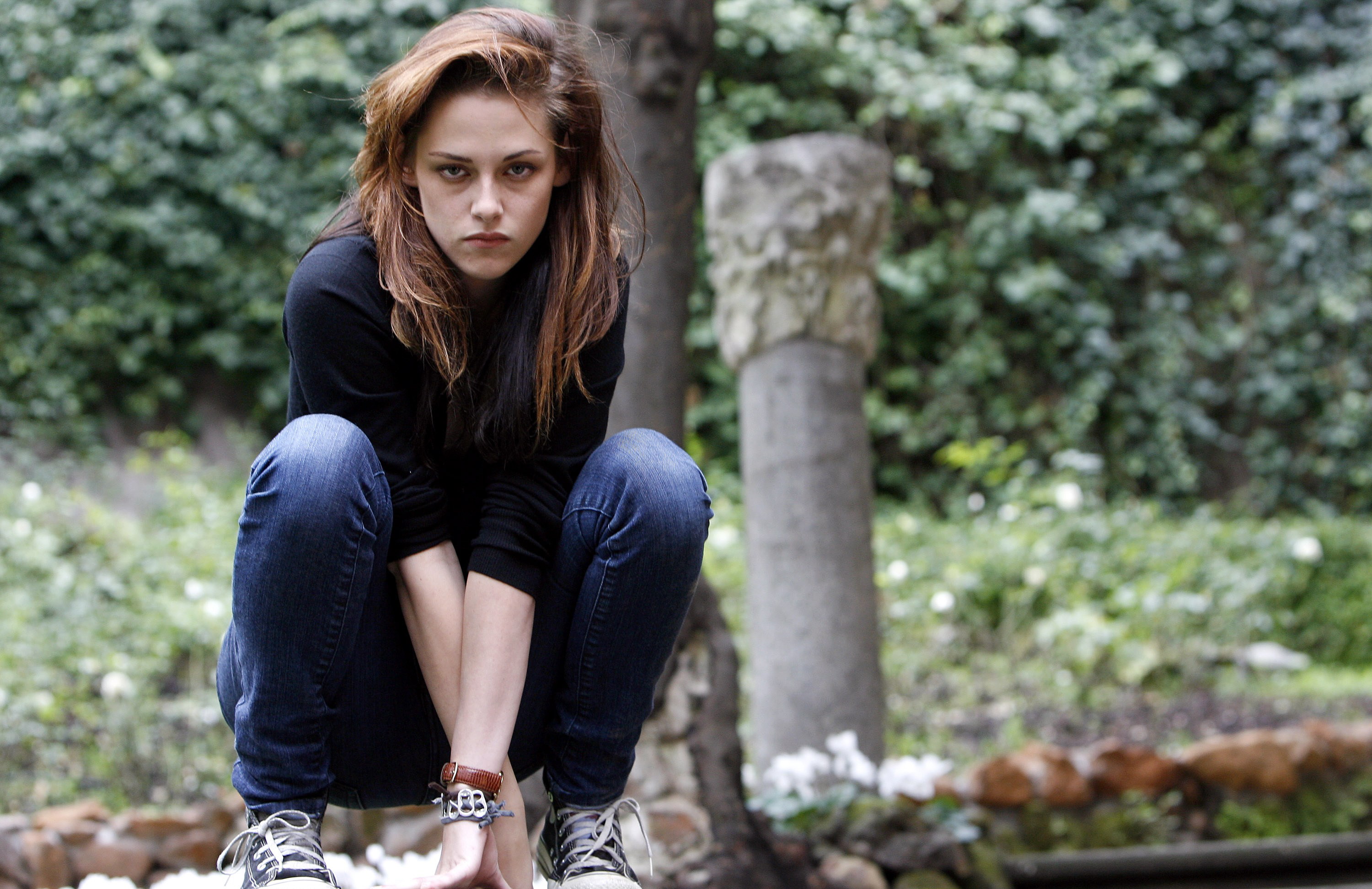 Kristen Stewart squatting on a wall in jeans and long sleeve black pullover